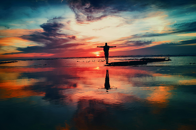 A man standing on the side of the ocean with arms raised, with a sunset in the foreground.